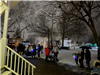 A group of diverse people are standing outside on the snow covered ground, in the background are illuminated holiday decorations