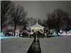 A white gazebo surrounded by snow on the ground, residents and illuminated holiday decorations