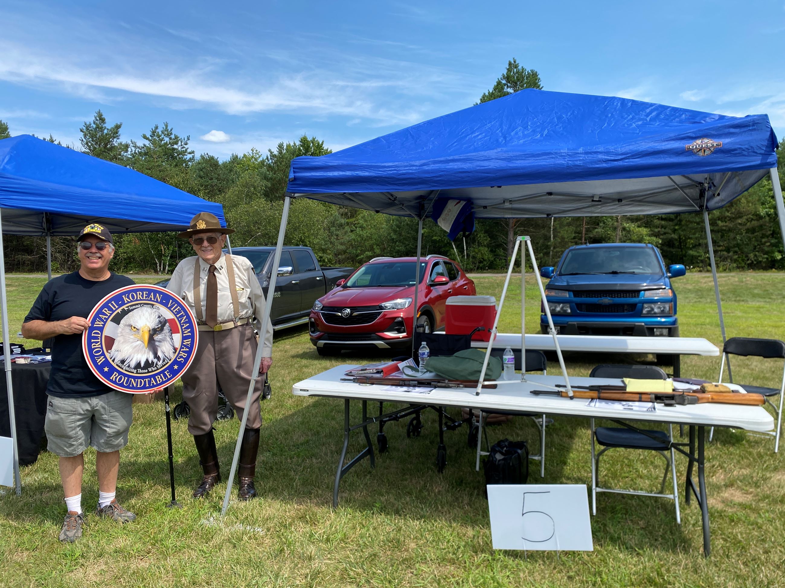 Vendor Set Up At Their Table Holding a Circular Sign with an Eagle in the Center at Copley Community Park on Saturday For Heritage Days 2025