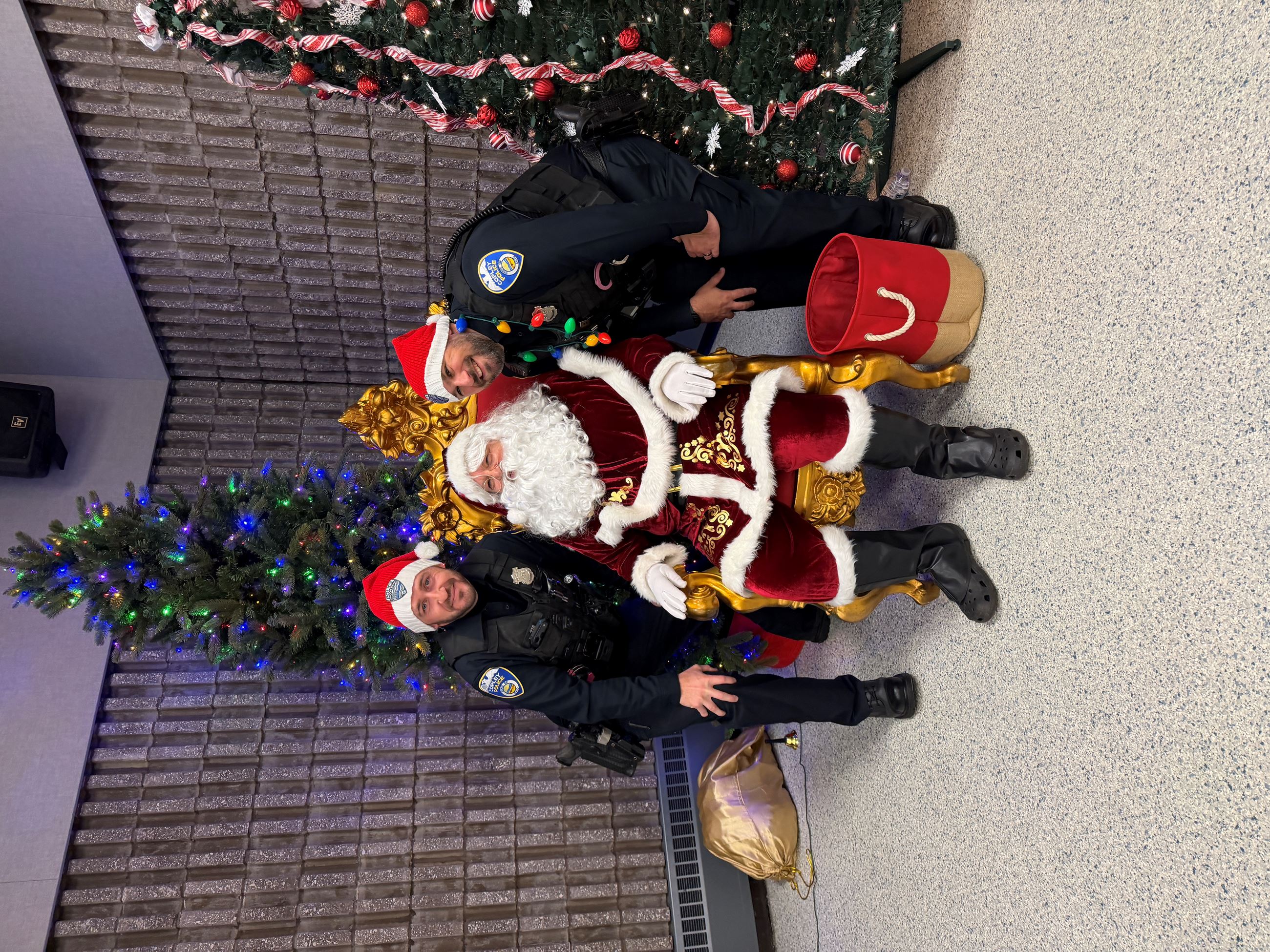 An officer Stands on either side of a sitting santa claus, between 2 Christmas trees