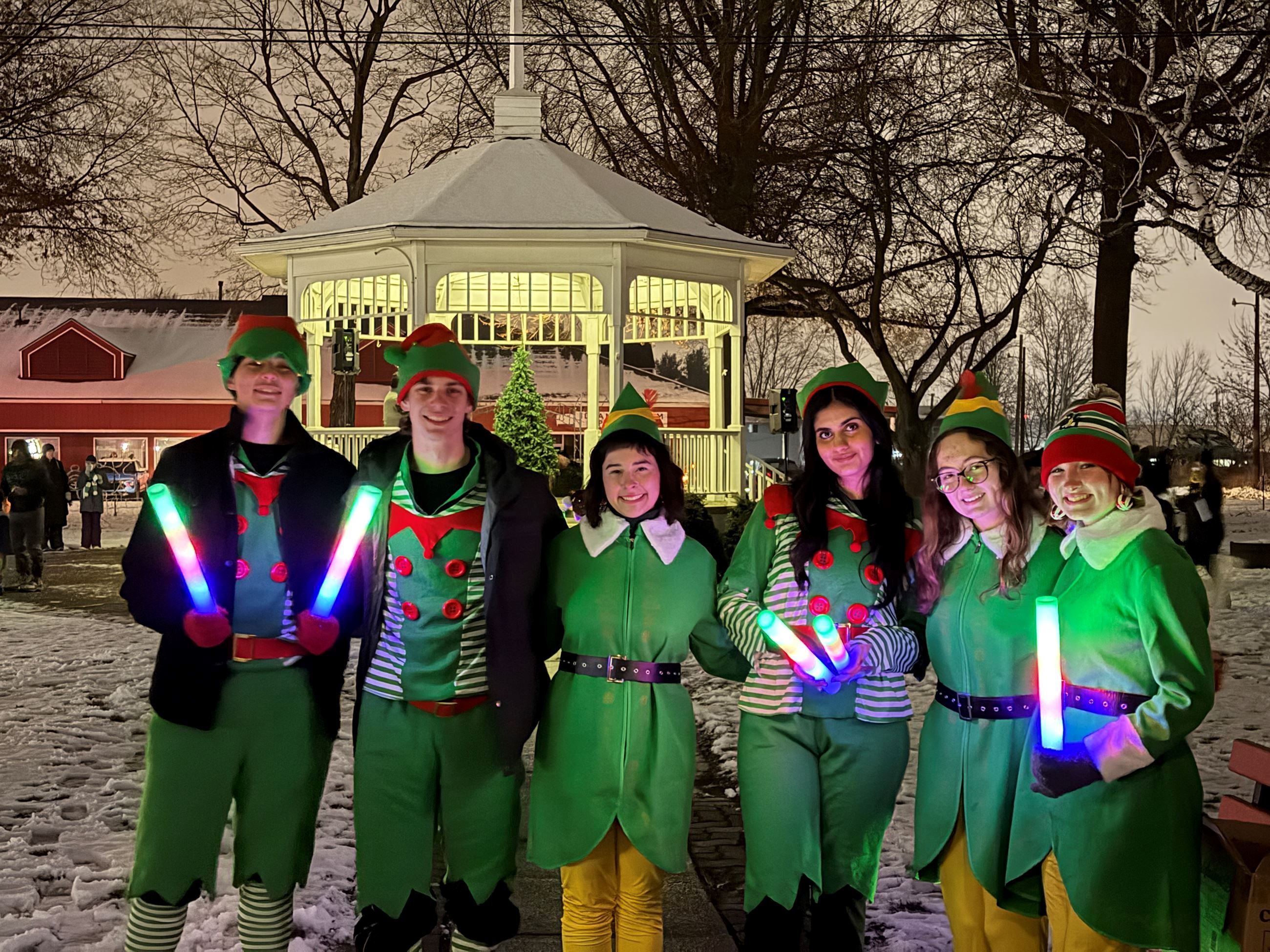 A group of high school aged kids are dressed in elf costumes holding light up wands, in the background is snow on the ground and a white gazebo