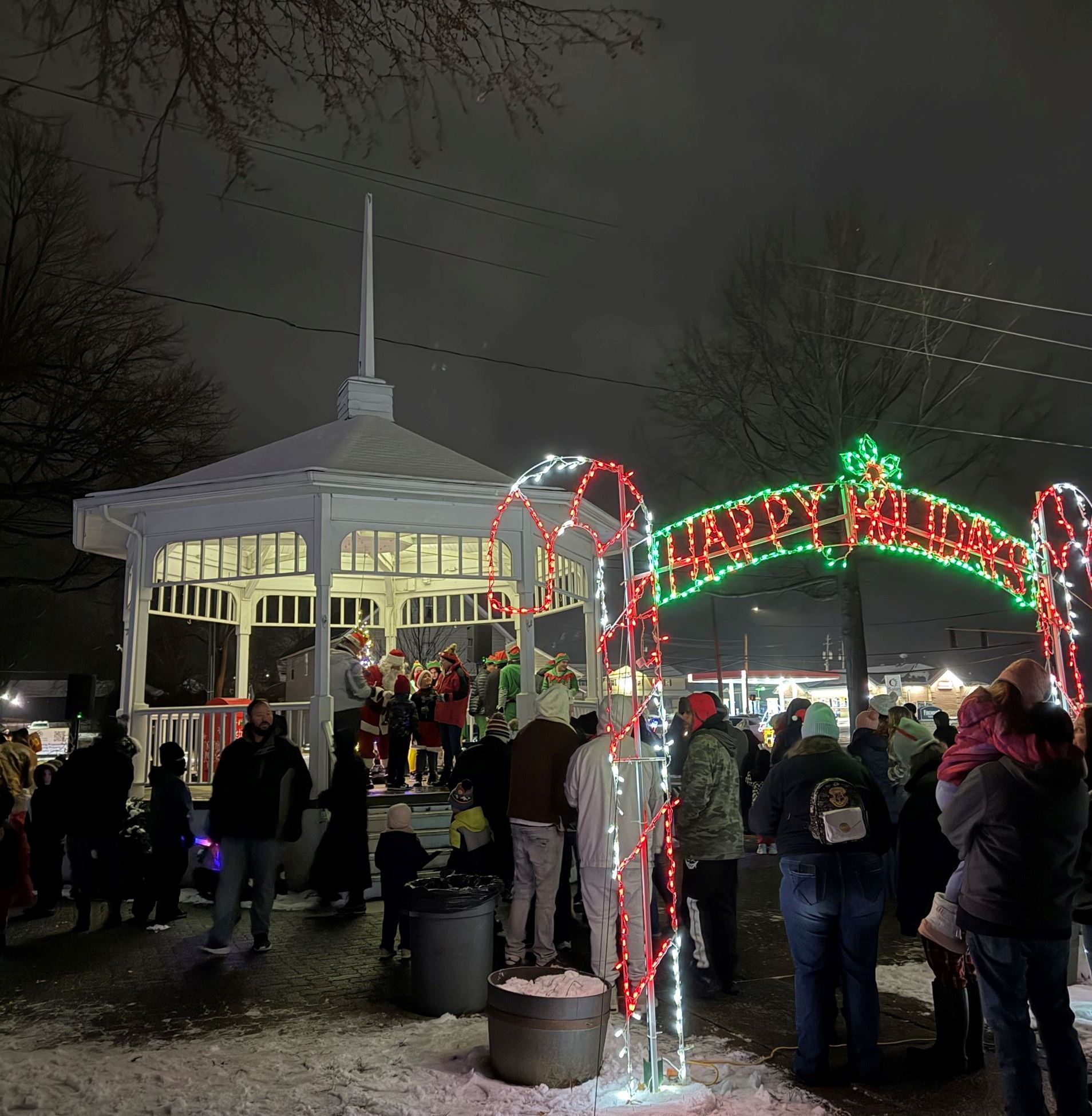 A group of people line up outside on the snow covered ground, the line goes under an illuminated holiday sign saying "Happy Holidays" the line goes towards a white gazebo where santa claus is taking pictures with children
