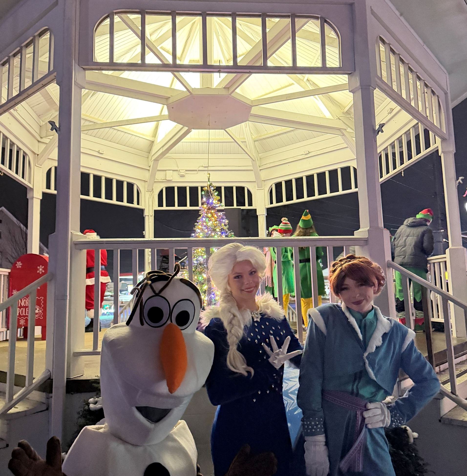 Olaf, Elsa and Anna pose for a photo together in front of the White Gazebo with an illuminated Christmas tree in the background