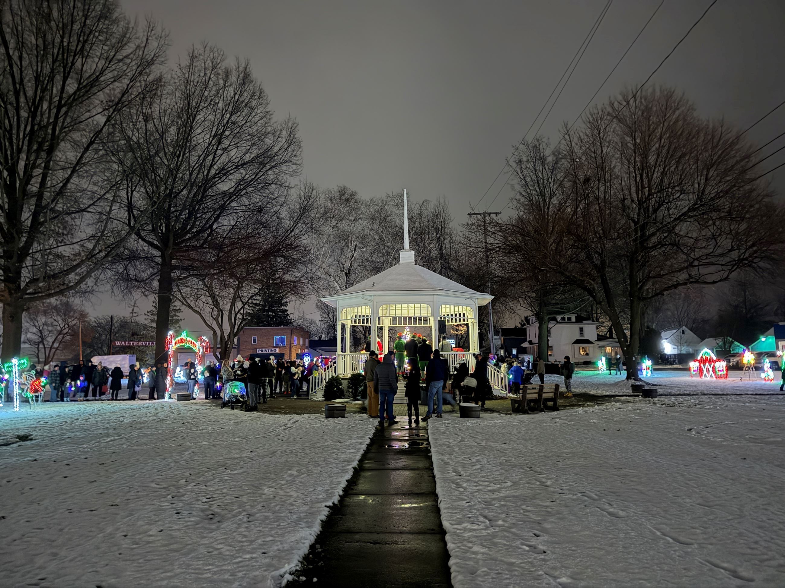 A white gazebo surrounded by snow on the ground, residents and illuminated holiday decorations